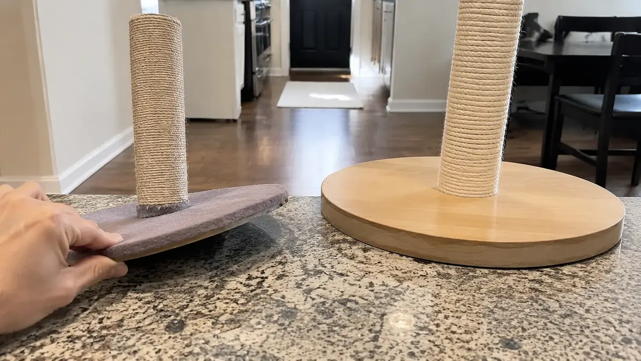 A person's hand pressing on the corner edge of a thin round cardboard-and-wood post base, showing it tilting and wobbling on a hardwood floor, a sturdy wide wood base nearby.